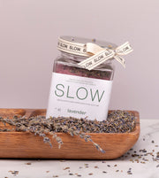 Esfoliante corporal emulsionado de açúcar e óleo essencial de alfazema, da SLOW, feito à mão em Portugal, num prato de madeira e sementes de alfazema, lavanda