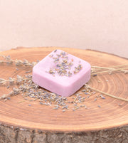 Loção hidratante sólida de alfazema, com óleo de coco e rosa mosqueta, da SLOW, feito à mão em Portugal, em cima de uma tábua de madeira e com sementes de aflazema, lavanda